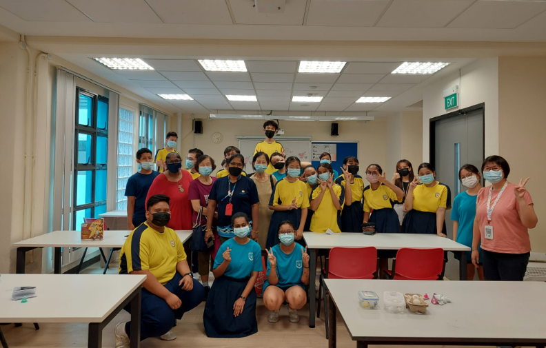 A group of students and teachers wearing face masks pose together in a classroom wearing yellow and blue uniforms.